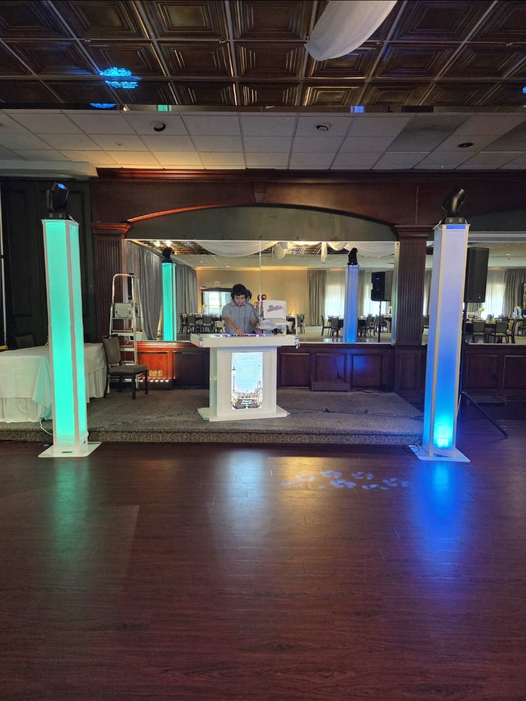 DJ Aiden Jules performing at a 50th anniversary celebration behind a white booth with tall teal and blue LED light towers in an elegant ballroom