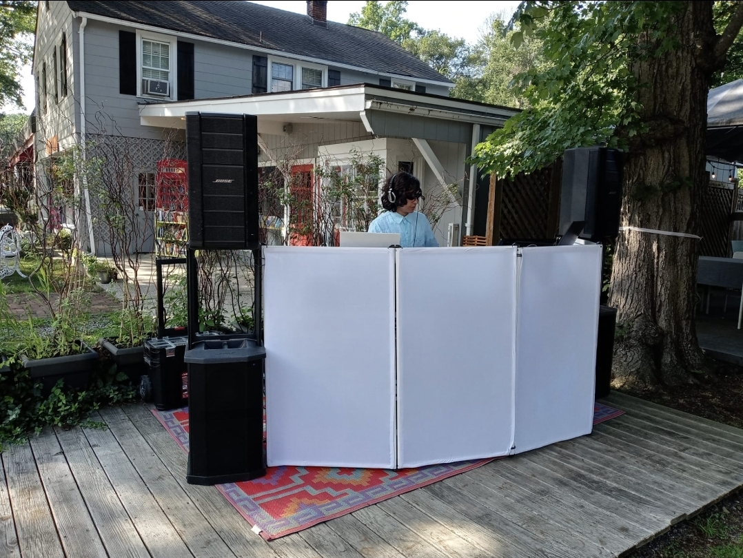 DJ Aiden Jules performing behind a white event booth with Bose speakers at an outdoor community gathering